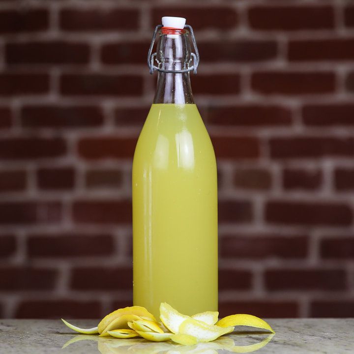 Láhev s houpačkou sedí na mramorové desce před cihlovou zdí. Láhev je naplněna zářivě žlutou tekutinou a před ní je seskupeno několik citronových slupek.
