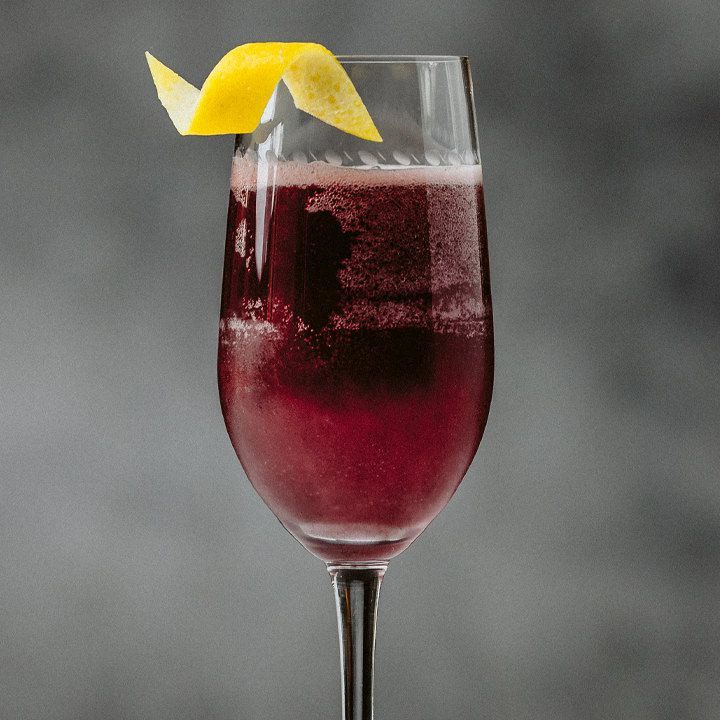 A taça de uma taça de champanhe é mostrada em close-up. É preenchido com uma bebida violeta borbulhante e decorado com um pequeno toque de limão enrolado. O fundo é cinza e difuso.