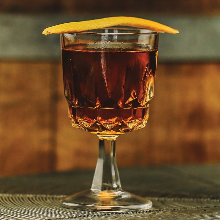 Um copo de coquetel atarracado e multifacetado com haste curta contém uma bebida em tons de cobre. Uma grande casca de laranja repousa sobre a boca do copo. O fundo é painéis de madeira, fora de foco.