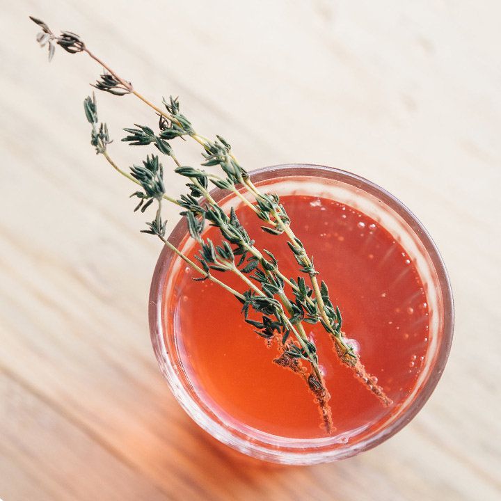 Un'inquadratura dall'alto mostra un bicchiere highball appoggiato su una superficie di legno duro chiaro. Il bicchiere è riempito con una bevanda frizzante rosso-arancio e guarnito con due rametti di timo.