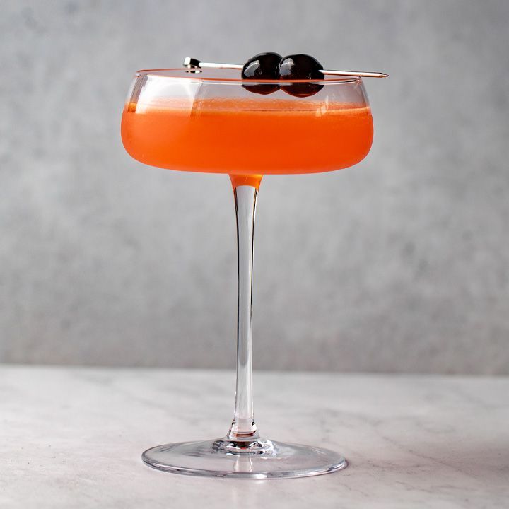 Un cupé de tija llarga amb un bol pla curt té un còctel de color taronja brillant. La beguda està guarnida amb dues cireres fosques sobre un pic de metall. El fons i el terra són de marbre gris.
