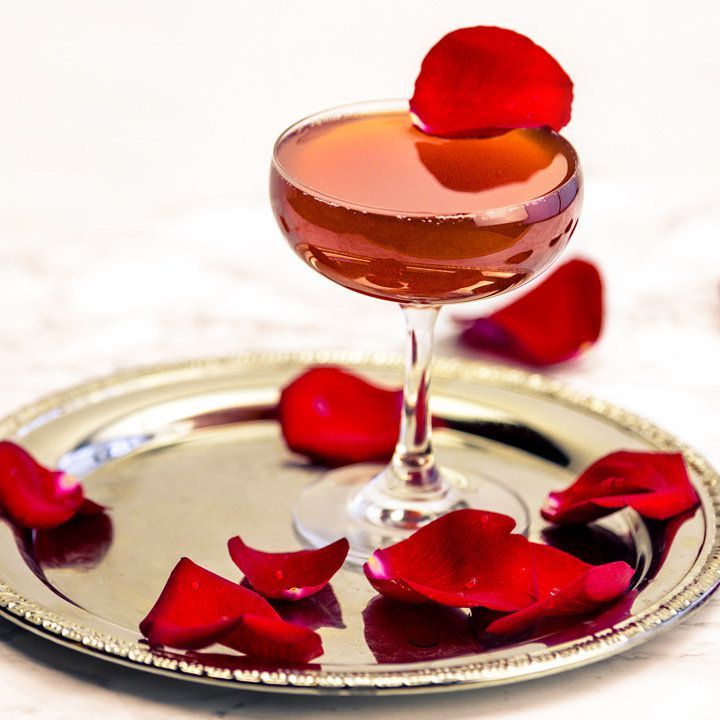 Uma taça de cupê está cheia até a borda com uma bebida rosa cintilante. O copo é coberto com uma única pétala de rosa vermelha, e muitas outras ficam em volta dela, em uma travessa dourada.