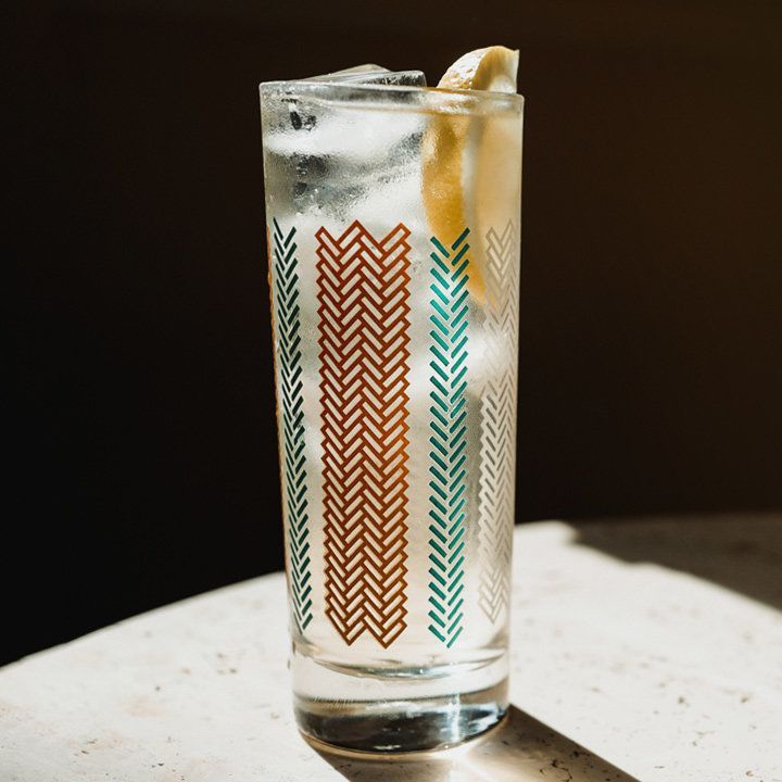 Un bicchiere highball si trova su una superficie di pietra illuminata in modo drammatico. È contrassegnato da chevron rossi e blu ed è riempito con una bevanda frizzante chiara, ghiaccio e uno spicchio di limone.